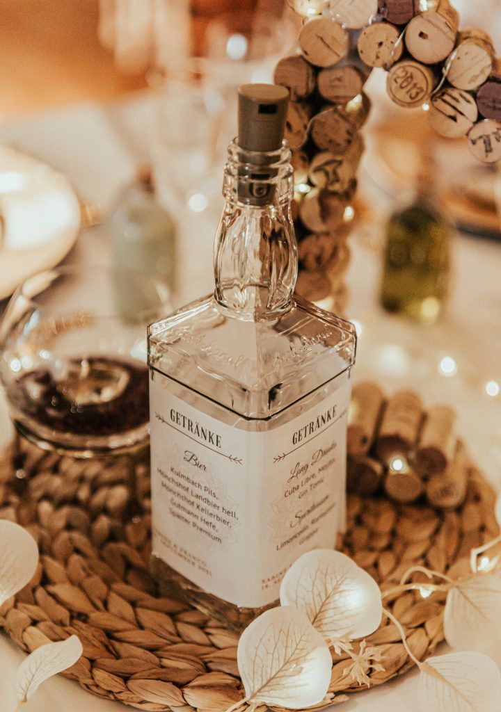 Deko mieten: Tischdekoration: leere Jack Daniel's Whiskey Flasche mit einer Drahtlichterkette, umwickelt mit der Getränkekarte auf Transparantpapier im Hochzeiitsdesign auf einem Flecht-Tischset