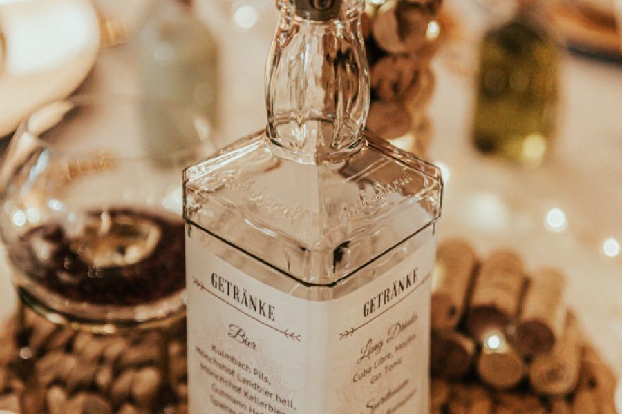 Tischdekoration: leere Jack Daniel's Whiskey Flasche mit einer Drahtlichterkette, umwickelt mit der Getränkekarte auf Transparantpapier im Hochzeiitsdesign auf einem Flecht-Tischset