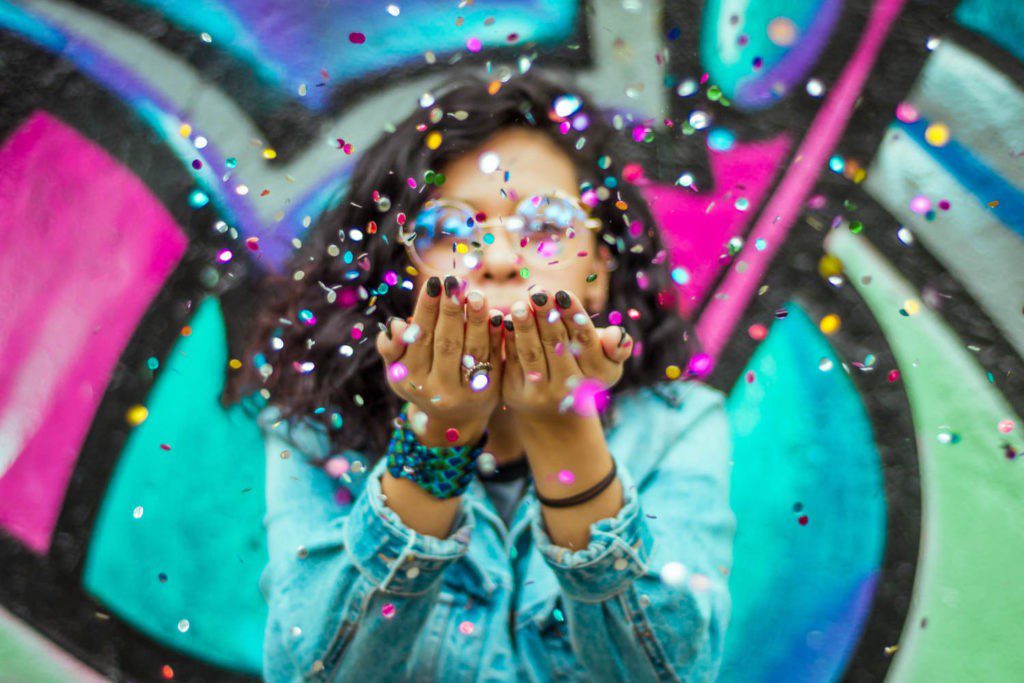Junge Frau in einer Jeansjacke, die vor einer bunt mit Graffiti besprühten Wand, pustet buntes Konfetti in die Luft
